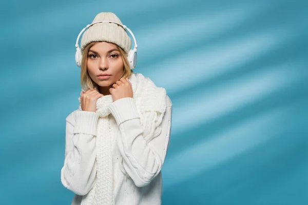 Young blonde woman in winter hat and wireless headphones listening music on blue background — Stock Photo