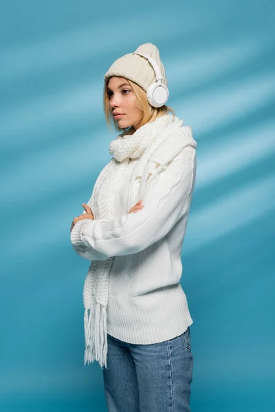 Mujer rubia joven en sombrero de invierno y auriculares inalámbricos escuchando música y de pie con los brazos cruzados en azul - foto de stock