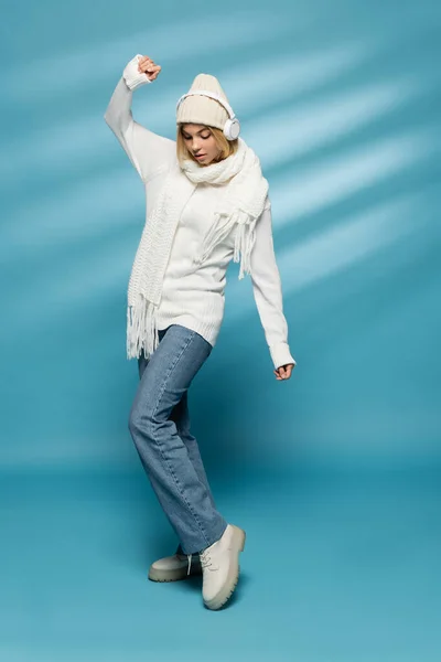 Longitud completa de mujer rubia joven en sombrero de invierno y auriculares inalámbricos posando en azul - foto de stock