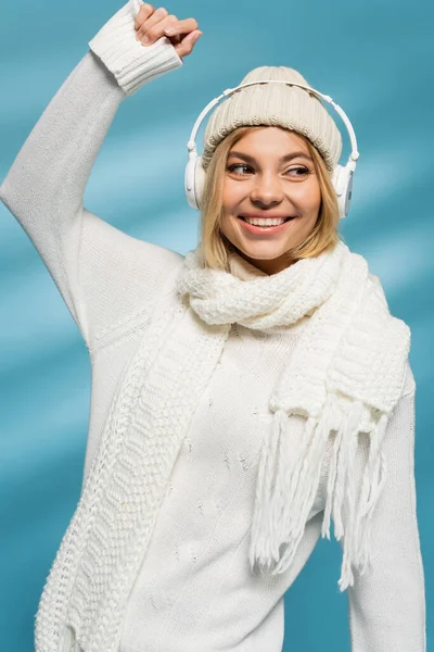 Happy blonde woman in winter hat and wireless headphones listening music on blue — Stock Photo