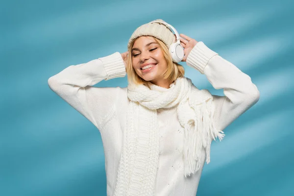 Mujer alegre en sombrero de invierno y auriculares inalámbricos escuchando música en azul - foto de stock