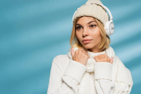 Blonde young woman in knitted hat and wireless headphones looking away on blue — Stock Photo
