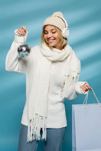 Heureux jeune femme en bonnet tricoté et écouteurs sans fil tenant boule disco avec sac à provisions sur bleu — Photo de stock