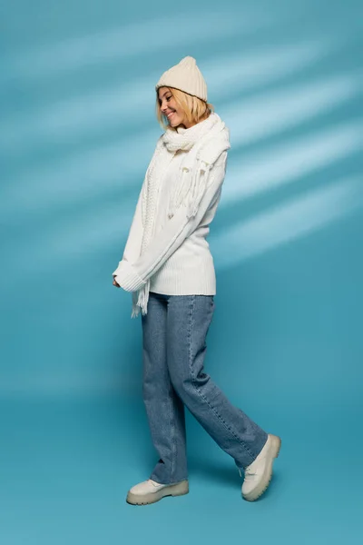 Full length of happy young woman in knitted hat and jeans smiling on blue — Stock Photo
