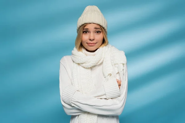 Pretty young woman in knitted hat and winter sweater feeling cold while standing with crossed arms on blue — Stock Photo