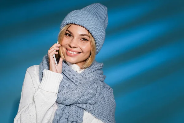 Alegre joven mujer en invierno sombrero y suéter hablando en el teléfono inteligente sobre fondo azul - foto de stock