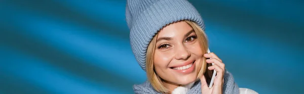 Cheerful young woman in winter hat and scarf talking on smartphone on blue background, banner — Stock Photo