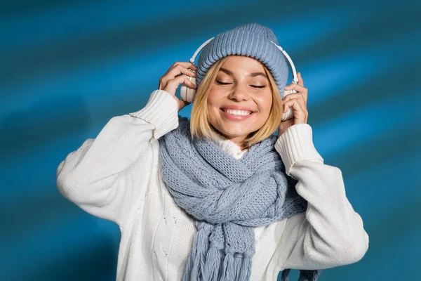 Cheerful young woman in winter scarf and hat wearing wireless headphones on blue — Stock Photo