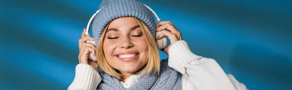 Fröhliche junge Frau in Winterschal und Mütze mit kabellosen Kopfhörern auf blau, Banner — Stockfoto