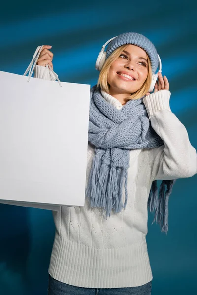 Allegra giovane donna in inverno sciarpa e cappello ascoltare musica in cuffie senza fili e tenendo shopping bag su blu — Foto stock