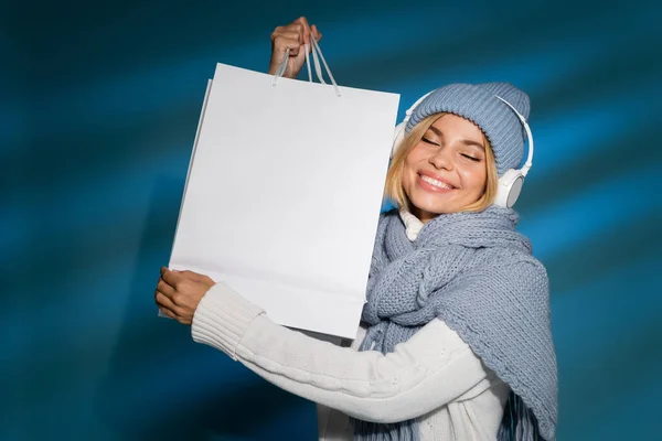 Heureux jeune femme en écharpe d'hiver et chapeau écouter de la musique dans les écouteurs sans fil et tenant sac à provisions sur bleu — Photo de stock