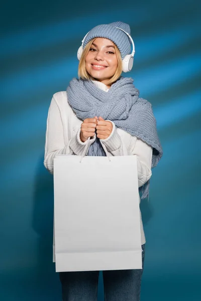 Femme joyeuse en chapeau d'hiver écouter de la musique dans des écouteurs sans fil et tenant sac à provisions sur bleu — Photo de stock