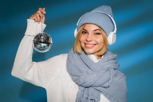 Donna allegra in cappello invernale e cuffie senza fili che tengono palla disco su blu — Foto stock