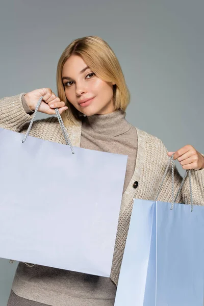 Blondine in Rollkragen und Strickjacke hält Einkaufstüten isoliert auf grau — Stockfoto