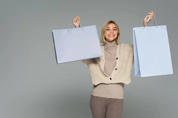 Happy blonde woman in turtleneck and cardigan holding shopping bags isolated on grey — Stock Photo