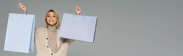 Mulher loira feliz em gola alta e casaco de lã segurando sacos de compras isolados em cinza, banner — Fotografia de Stock