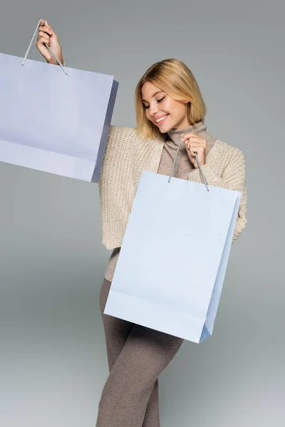Pleased blonde woman in turtleneck and cardigan holding shopping bags on grey — Stock Photo