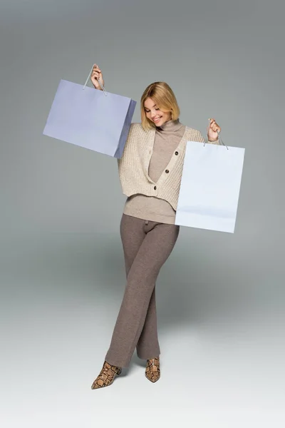На всю длину счастливой женщины в трикотажных штанах и кардигане, держащей сумки с покупками на сером — стоковое фото