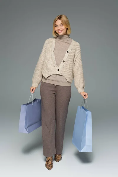 Pleine longueur de femme gaie en pantalon tricoté et cardigan tenant des sacs à provisions sur gris — Photo de stock