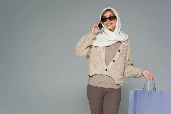 Mujer alegre en prendas de punto y gafas de sol sosteniendo bolsas de compras mientras habla en el teléfono inteligente aislado en gris - foto de stock