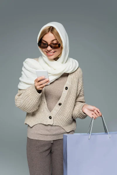 Mujer alegre en prendas de punto y gafas de sol sosteniendo bolsas de compras durante el uso de teléfono inteligente aislado en gris - foto de stock