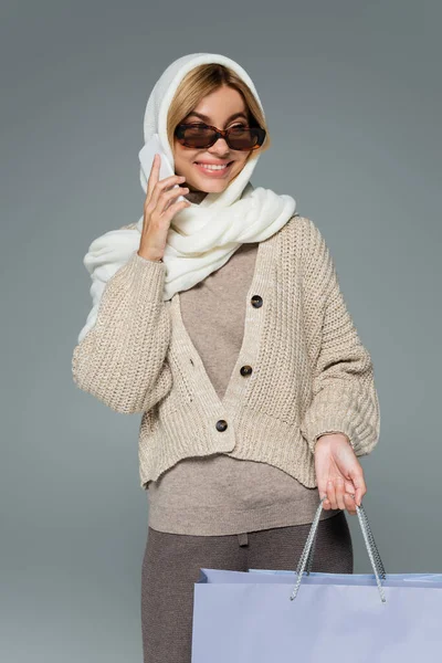 Mulher alegre em malhas e óculos de sol segurando sacos de compras enquanto conversa no smartphone isolado em cinza — Fotografia de Stock
