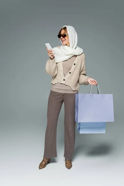 Full length of cheerful woman in knitwear and sunglasses holding shopping bags while using smartphone on grey — Stock Photo
