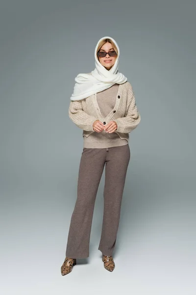 Pleine longueur de femme gaie en tricot et des lunettes de soleil élégantes debout sur le gris — Photo de stock