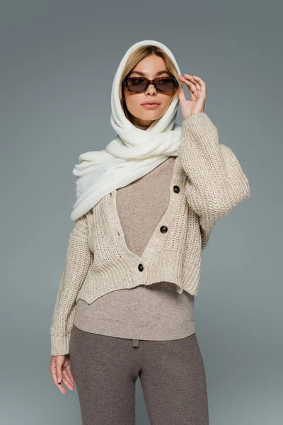 Jeune femme en bonneterie et foulard ajustant les lunettes de soleil isolées sur gris — Photo de stock
