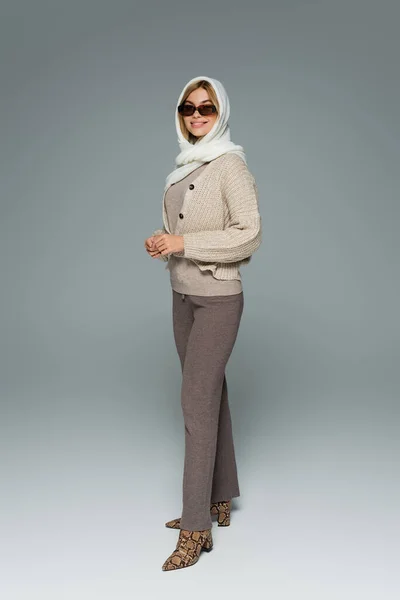 Longitud completa de la mujer feliz en pañuelo para la cabeza y gafas de sol elegantes posando en gris - foto de stock
