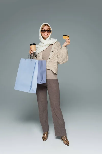 Pleine longueur de femme heureuse dans des lunettes de soleil tenant tasse en papier et carte de crédit tout en se tenant debout avec des sacs à provisions sur gris — Photo de stock