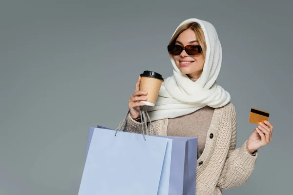 Mujer feliz en gafas de sol sosteniendo taza de papel y tarjeta de crédito mientras está de pie con bolsas aisladas en gris - foto de stock