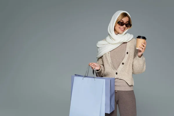 Cheerful woman in winter headscarf and sunglasses holding paper cup and shopping bags isolated on grey — Stock Photo