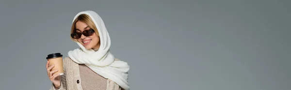 Donna allegra in inverno velo e occhiali da sole tenendo tazza di carta isolata su grigio, banner — Foto stock