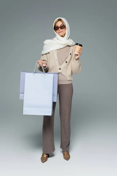 Longitud completa de la mujer alegre en invierno pañuelo para la cabeza y gafas de sol sosteniendo taza de papel y bolsas de compras en gris - foto de stock