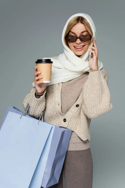 Веселая женщина в солнечных очках, держа бумажную чашку и сумки для покупок, разговаривая на смартфоне, изолированном на сером — стоковое фото
