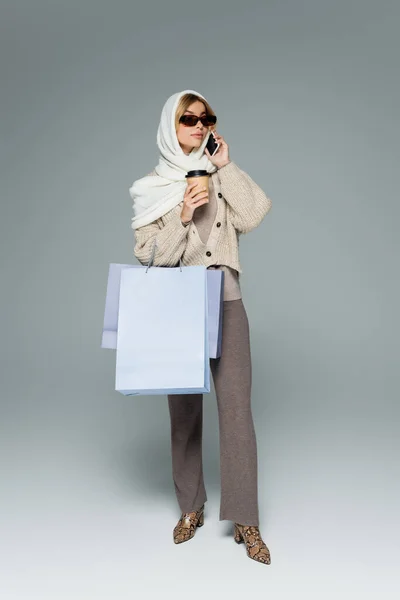 Полная длина женщины в платке и солнцезащитных очках с бумажной чашкой и пакетами для покупок во время разговора по смартфону на сером — стоковое фото