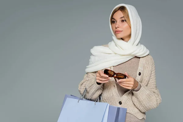 Young woman in headscarf holding stylish sunglasses and shopping bags isolated on grey — Stock Photo