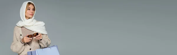 Giovane donna in inverno velo che tiene occhiali da sole alla moda e borse della spesa isolate su grigio, banner — Foto stock