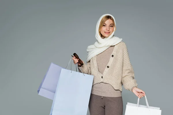 Mujer joven complacida en invierno pañuelo con gafas de sol elegantes y bolsas de compras aisladas en gris - foto de stock