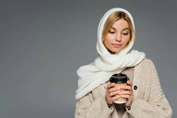 Mujer joven en invierno pañuelo en la cabeza sosteniendo taza de papel con bebida para llevar aislado en gris - foto de stock