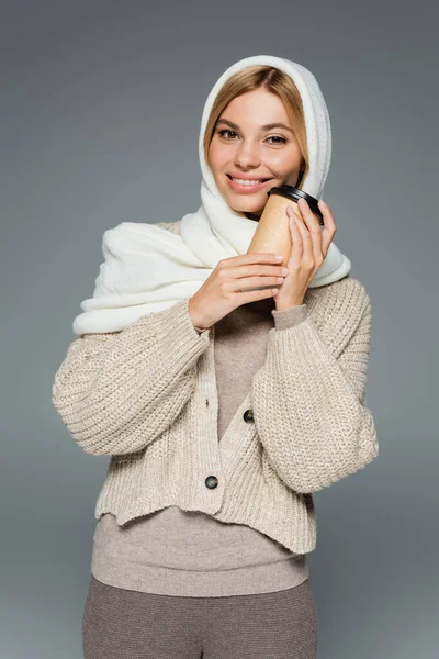 Fröhliche junge Frau im winterlichen Kopftuch hält Pappbecher mit Imbissgetränk isoliert auf grau — Stockfoto