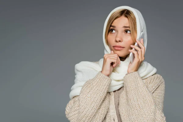 Mujer joven preocupada en el pañuelo de cabeza de invierno hablando en el teléfono móvil y mirando hacia otro lado aislado en gris - foto de stock