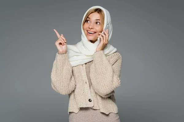 Fröhliche junge Frau im winterlichen Kopftuch telefoniert mit Handy und zeigt mit dem Finger isoliert auf grau — Stockfoto