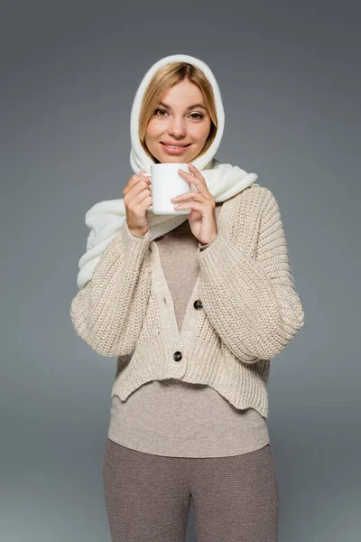 Mujer feliz en invierno pañuelo para la cabeza y punto cárdigan celebración taza de café aislado en gris - foto de stock