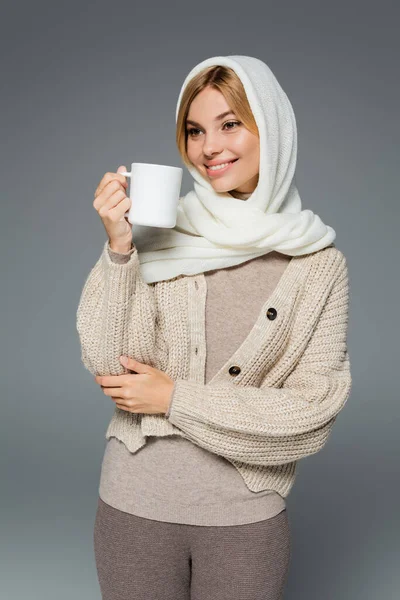 Mujer alegre en invierno pañuelo para la cabeza y cárdigan celebración taza de café aislado en gris - foto de stock