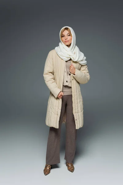 Повна довжина стильної молодої жінки в зимових головних уборах і пальто, стоячи на сірому — Stock Photo