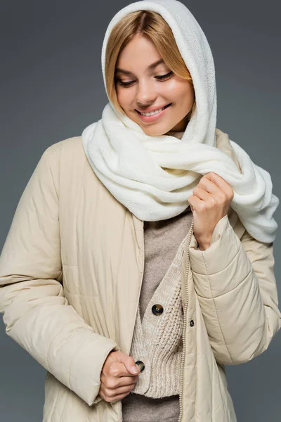 Позитивная молодая женщина в зимнем платке регулируя пиджак, изолированный на сером — стоковое фото