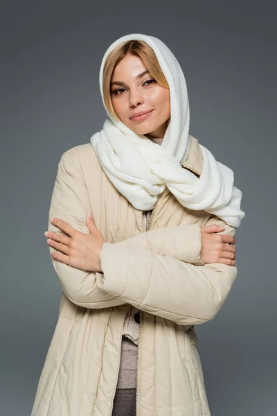 Mujer joven con estilo en invierno pañuelo para la cabeza y chaqueta hinchable de pie con brazos cruzados aislados en gris - foto de stock