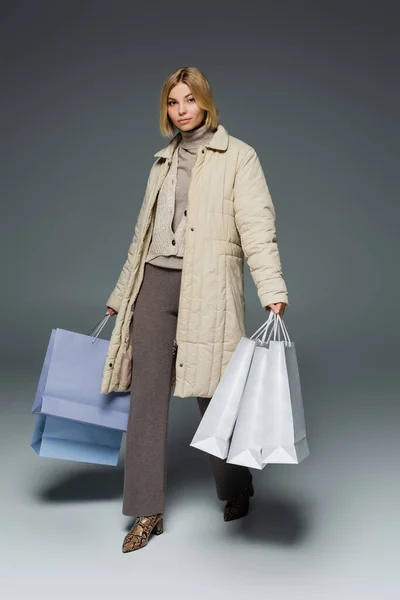 Full length of blonde young woman in puffer coat holding shopping bags while walking on grey — Stock Photo